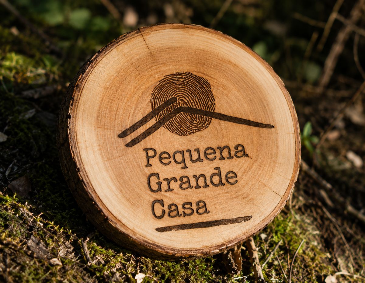 Logótipo da Pequena Grande Casa gravado em medalhão de madeira natural, sobre musgo da floresta